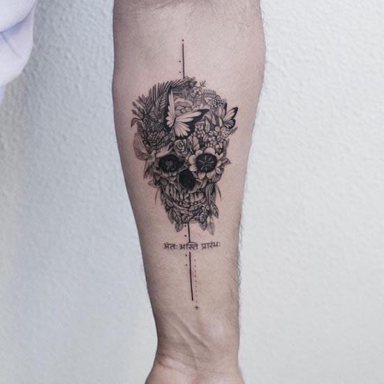 skull covered with flowers leaves butterfly sanskrit text at bottom tattoo male forearm