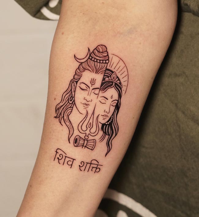 half shiva face half parvati face trishul damru in between below sanskrit words shiva shakti tattoo on female arm